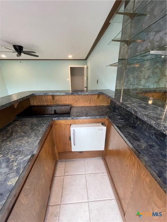 kitchen featuring brown cabinetry, dark stone counters, light tile patterned flooring, and ceiling fan