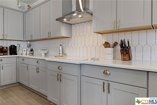 kitchen featuring light hardwood / wood-style floors, decorative backsplash, wall chimney exhaust hood, gray cabinets, and stovetop