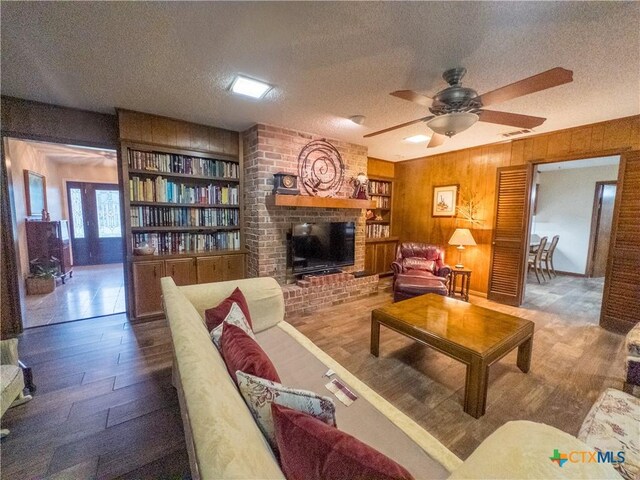 living room featuring a textured ceiling, a brick fireplace, wood finished floors, a ceiling fan, and wood walls