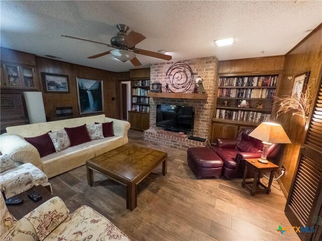 living room with wood walls, a ceiling fan, a textured ceiling, wood finished floors, and a brick fireplace