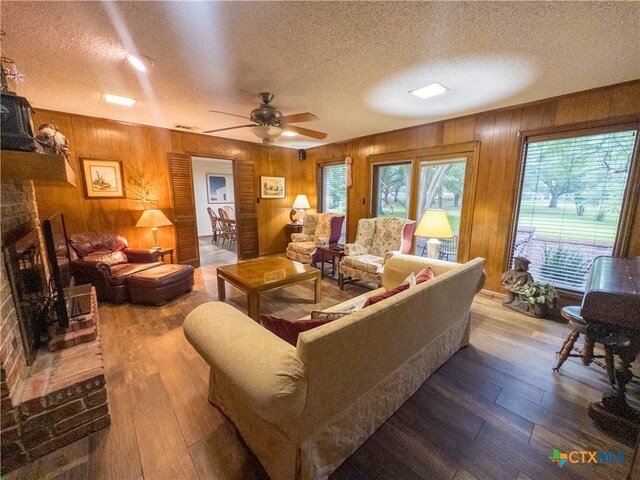 living room with a textured ceiling, wood walls, hardwood / wood-style flooring, a ceiling fan, and a brick fireplace