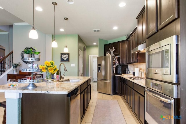 kitchen with an island with sink, stainless steel appliances, hanging light fixtures, light stone countertops, and sink