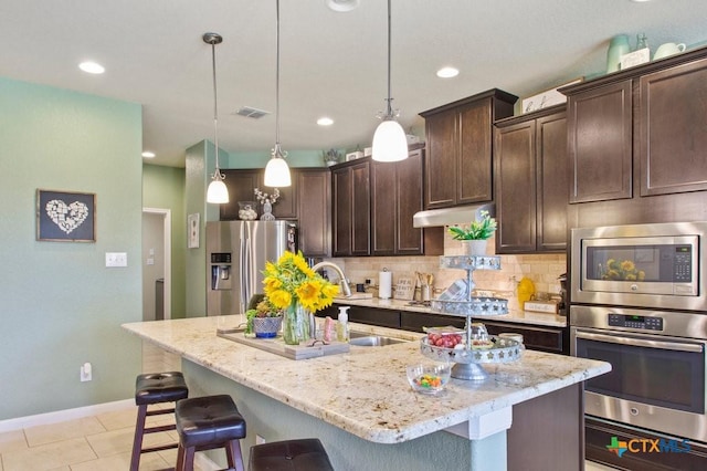 kitchen with light stone countertops, dark brown cabinetry, appliances with stainless steel finishes, decorative light fixtures, and a center island with sink