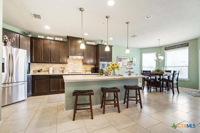 kitchen with dark brown cabinetry, an island with sink, hanging light fixtures, stainless steel fridge, and a breakfast bar area