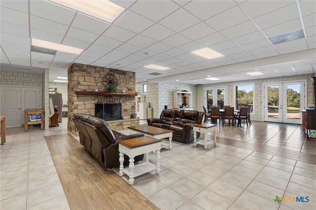 living room featuring a drop ceiling, a stone fireplace, light tile patterned floors, and french doors