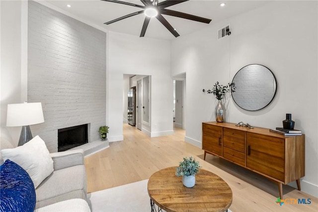 living area with a ceiling fan, light wood-style flooring, baseboards, a brick fireplace, and a high ceiling