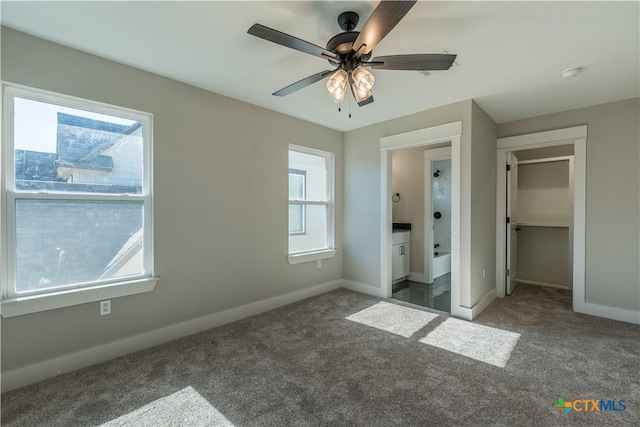 unfurnished bedroom with ensuite bathroom, a walk in closet, dark carpet, a closet, and ceiling fan