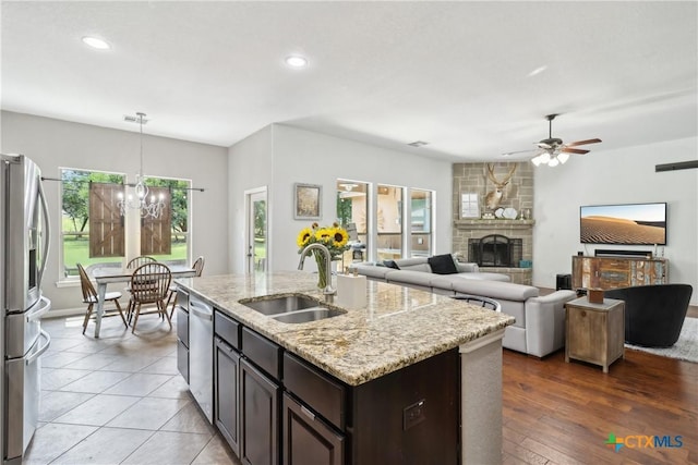 kitchen featuring stainless steel appliances, a sink, dark brown cabinets, ceiling fan with notable chandelier, and a fireplace