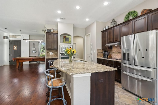 kitchen with arched walkways, a sink, light stone countertops, stainless steel refrigerator with ice dispenser, and under cabinet range hood