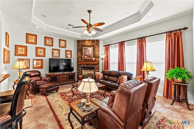 living room with a tray ceiling, crown molding, ceiling fan, and a brick fireplace