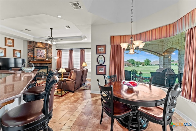 dining space featuring a fireplace, ceiling fan with notable chandelier, light tile patterned floors, and a tray ceiling