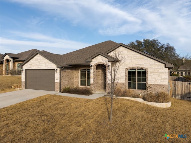 french country inspired facade featuring roof with shingles, concrete driveway, an attached garage, a front yard, and fence