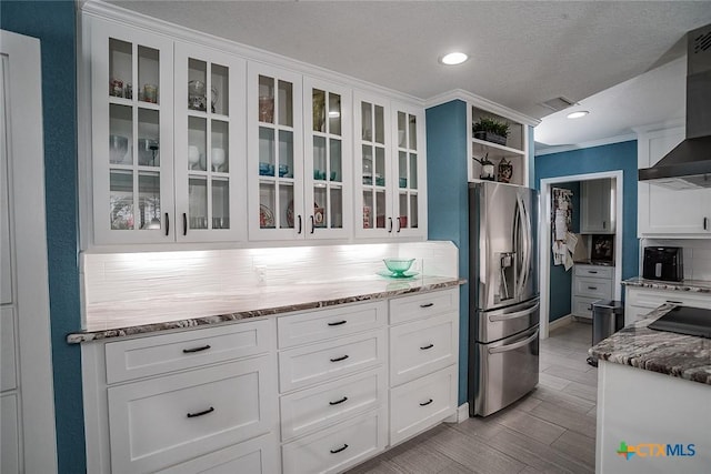 kitchen with light stone counters, white cabinetry, wall chimney exhaust hood, stainless steel fridge, and glass insert cabinets