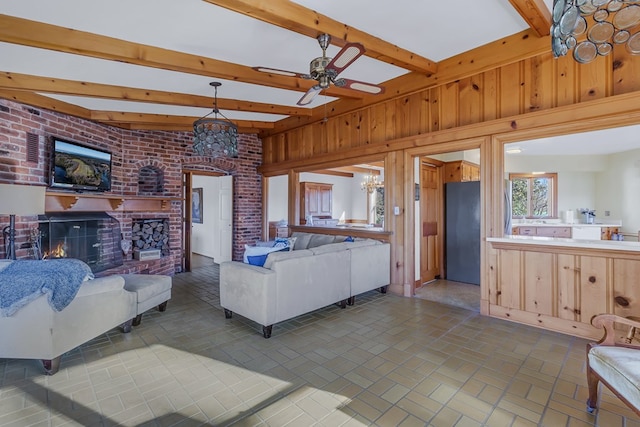 living room featuring ceiling fan with notable chandelier, beam ceiling, a fireplace, and wooden walls