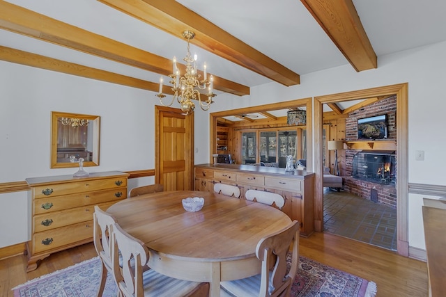 dining area featuring beam ceiling, light wood-type flooring, a notable chandelier, and a brick fireplace