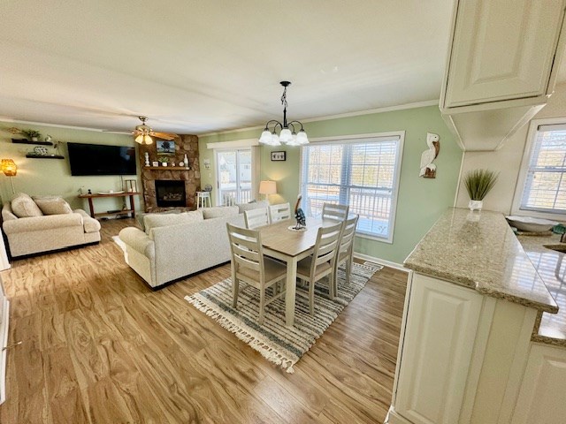 dining space featuring light wood-type flooring, ceiling fan with notable chandelier, crown molding, and a stone fireplace