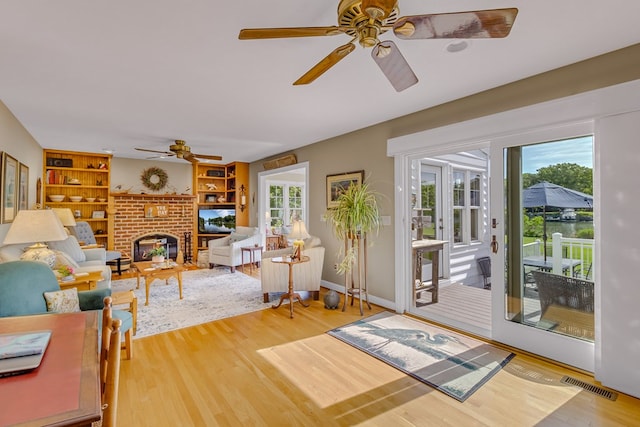 living room with ceiling fan, visible vents, a brick fireplace, built in shelves, and wood finished floors