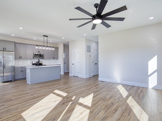 kitchen with light wood-type flooring, stainless steel appliances, decorative light fixtures, gray cabinets, and an island with sink