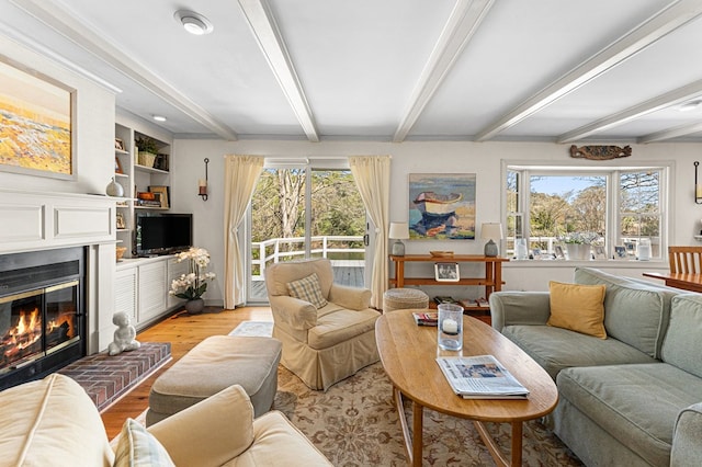 living area featuring beamed ceiling, a glass covered fireplace, plenty of natural light, and light wood-style floors
