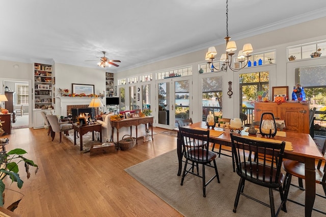 dining area featuring built in shelves, a brick fireplace, light hardwood / wood-style flooring, crown molding, and ceiling fan with notable chandelier