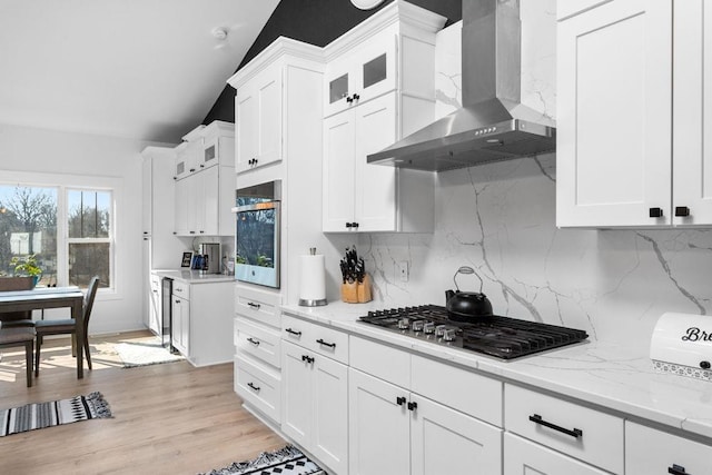 kitchen with wall chimney exhaust hood, stainless steel appliances, backsplash, white cabinets, and vaulted ceiling