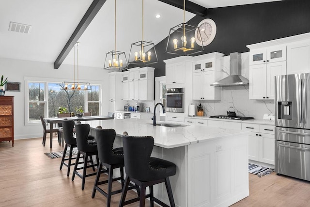 kitchen with decorative light fixtures, sink, wall chimney range hood, stainless steel appliances, and a center island with sink