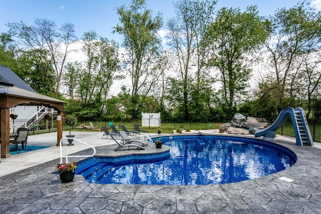 view of pool with a water slide, a patio area, fence, and view of wooded area