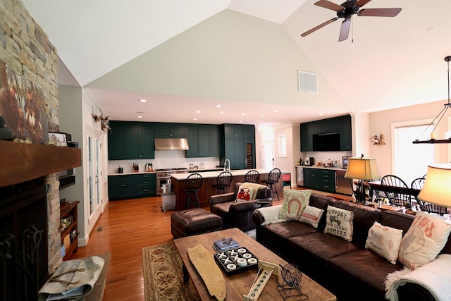 living room with ceiling fan, light hardwood / wood-style floors, a stone fireplace, and high vaulted ceiling