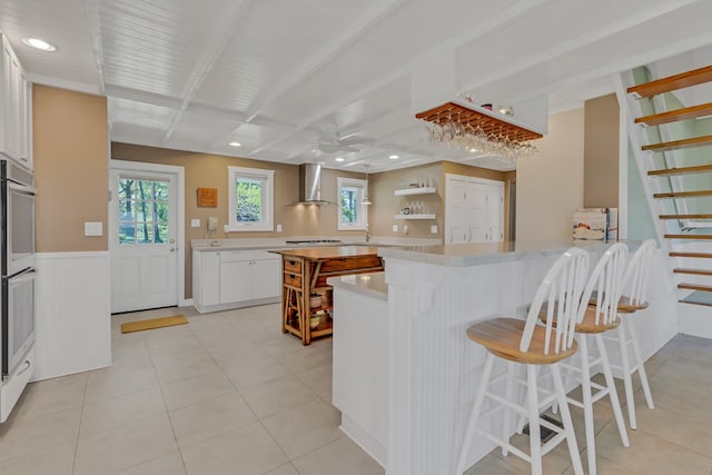 kitchen featuring light tile patterned flooring, a kitchen bar, white cabinetry, kitchen peninsula, and wall chimney range hood