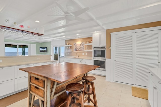 kitchen featuring sink, light tile patterned floors, stainless steel double oven, beam ceiling, and white cabinets