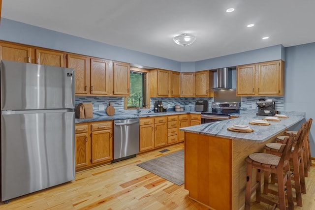 kitchen with wall chimney range hood, kitchen peninsula, light hardwood / wood-style floors, a kitchen bar, and appliances with stainless steel finishes