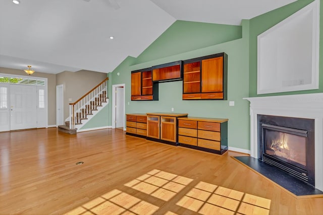 unfurnished living room featuring stairs, a glass covered fireplace, baseboards, high vaulted ceiling, and light wood-style flooring