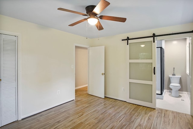 unfurnished bedroom with light hardwood / wood-style flooring, ceiling fan, a barn door, connected bathroom, and a closet