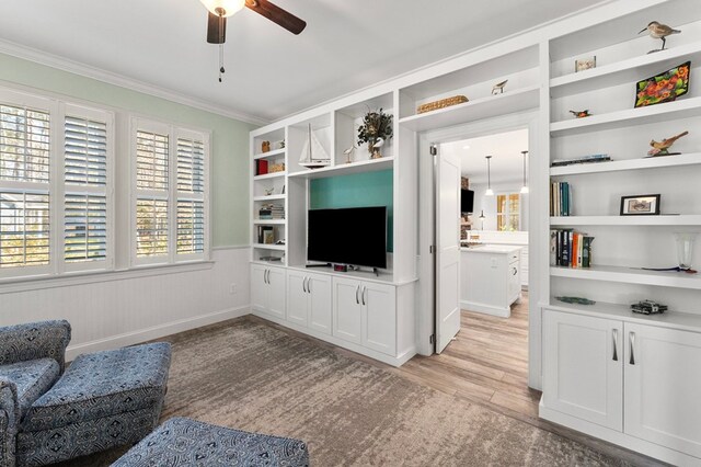 living area featuring built in shelves, a ceiling fan, a wainscoted wall, light wood-style flooring, and crown molding