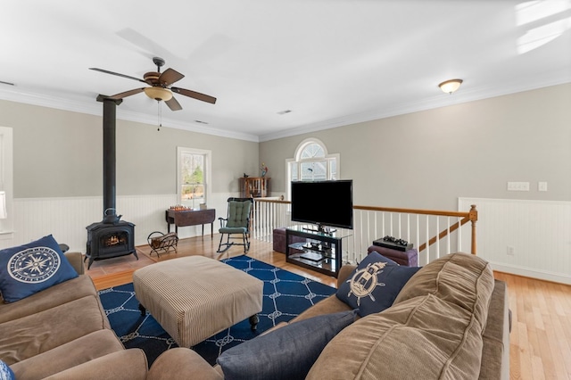 living room with wood finished floors, a wainscoted wall, a wood stove, ceiling fan, and ornamental molding