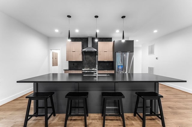 kitchen featuring pendant lighting, a center island with sink, wall chimney range hood, sink, and stainless steel fridge