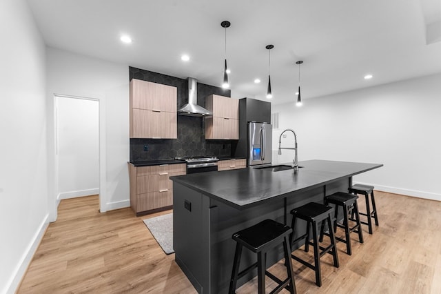 kitchen featuring wall chimney exhaust hood, stainless steel appliances, pendant lighting, a center island with sink, and light hardwood / wood-style floors