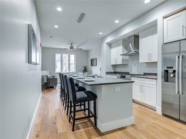 kitchen with a kitchen breakfast bar, ceiling fan, light hardwood / wood-style flooring, wall chimney exhaust hood, and stainless steel fridge