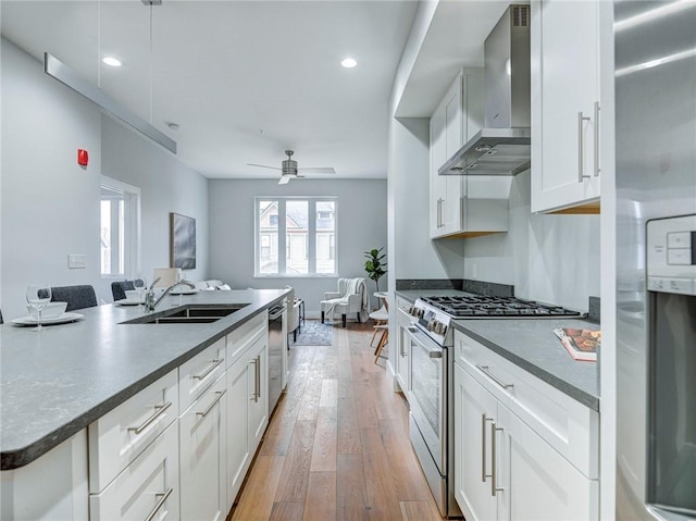 kitchen featuring appliances with stainless steel finishes, white cabinetry, light hardwood / wood-style flooring, ceiling fan, and wall chimney range hood