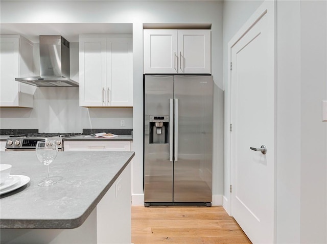 kitchen featuring wall chimney range hood, stove, stainless steel fridge with ice dispenser, white cabinets, and light wood-type flooring