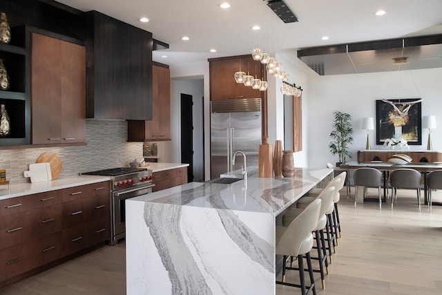 kitchen with tasteful backsplash, high end appliances, light stone counters, an island with sink, and decorative light fixtures