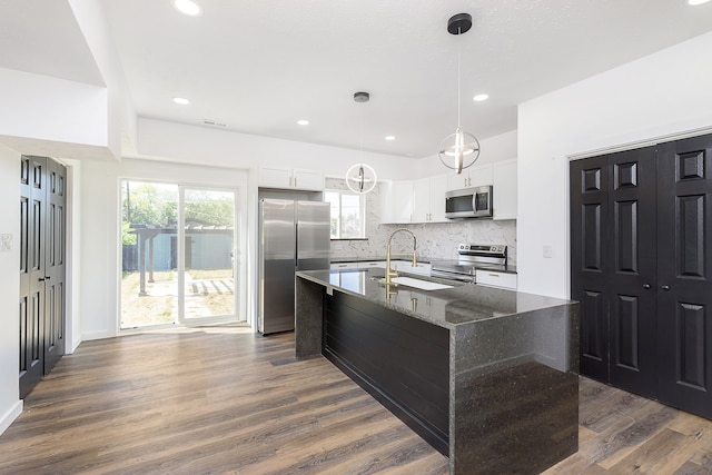 kitchen featuring white cabinetry, backsplash, appliances with stainless steel finishes, dark stone countertops, and a center island with sink