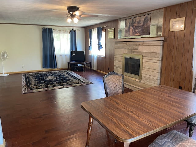 living room with dark wood-type flooring, wooden walls, ceiling fan, ornamental molding, and a stone fireplace