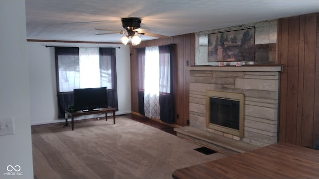 living room featuring wooden walls, carpet flooring, ceiling fan, and a stone fireplace