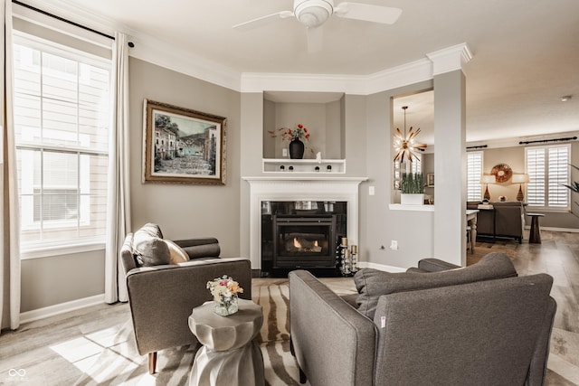 living area featuring crown molding, baseboards, ceiling fan with notable chandelier, light wood-style floors, and a glass covered fireplace