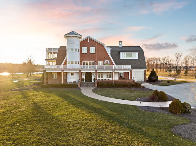 back of property at dusk featuring an attached garage, a balcony, a gambrel roof, driveway, and a lawn