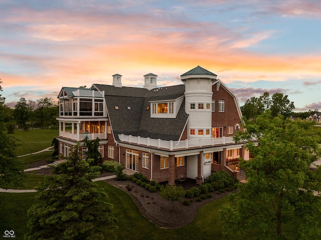 back of property with a balcony, brick siding, a lawn, and a gambrel roof