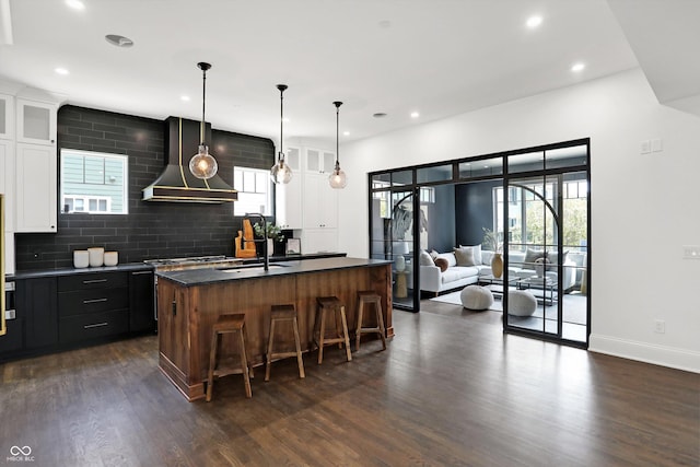 kitchen with white cabinetry, dark hardwood / wood-style floors, an island with sink, decorative light fixtures, and wall chimney exhaust hood