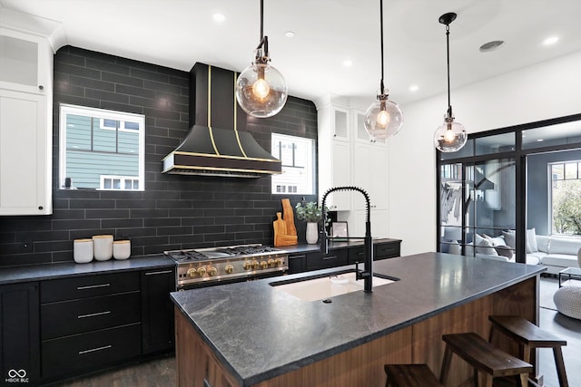 kitchen with tasteful backsplash, an island with sink, stove, wall chimney range hood, and white cabinets