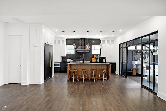 kitchen with wall chimney range hood, built in refrigerator, white cabinetry, a center island with sink, and a kitchen bar